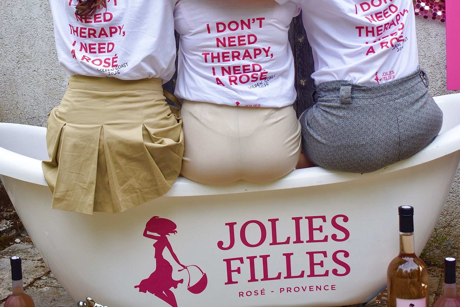 Drei Frauen sitzen auf einer Badewanne mit dem Schriftzug Jolies Filles, Rosé aus der Provence, daneben Flaschen des Weins