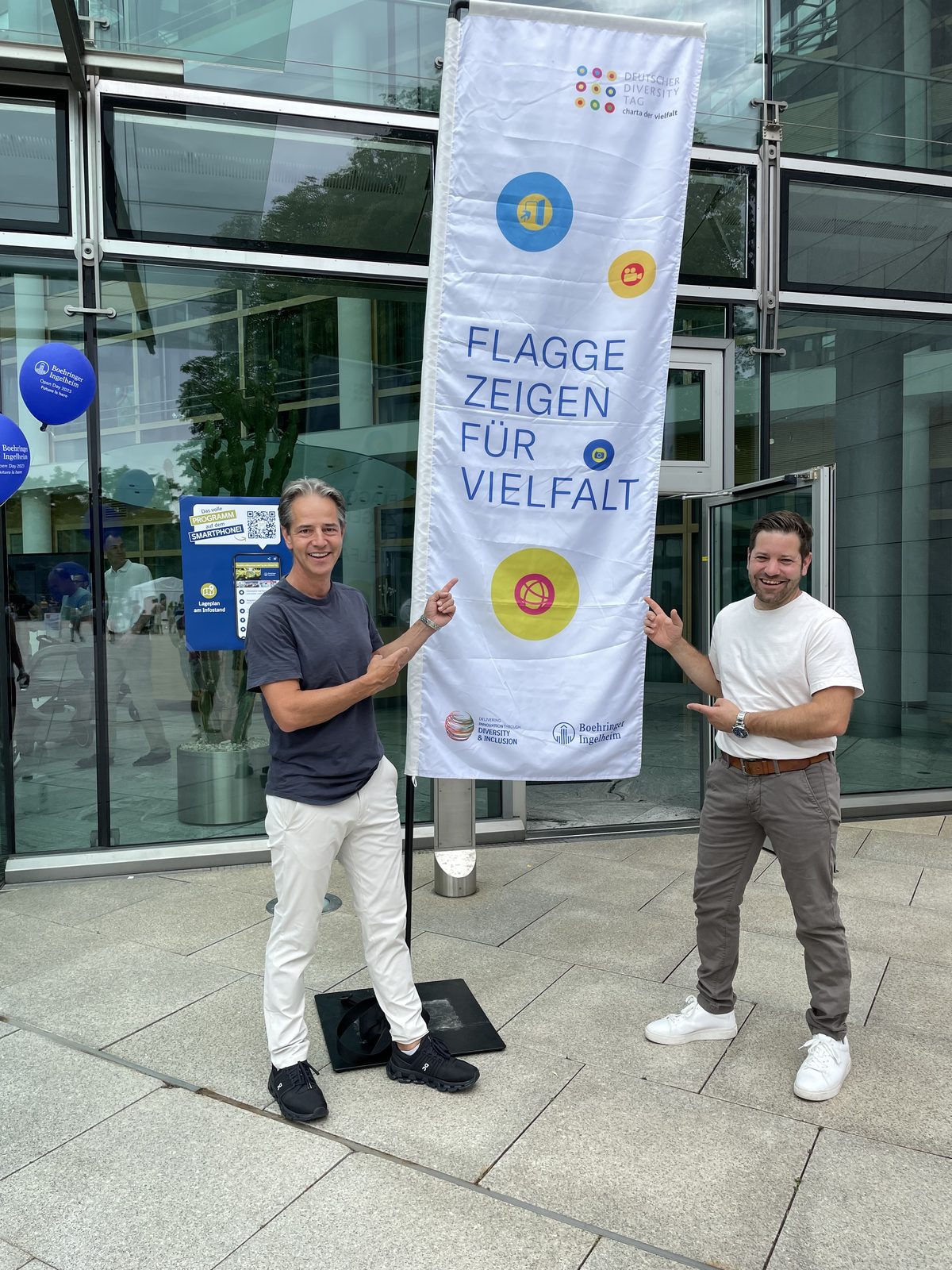 Zwei Männer vor einer Fahne mit der Aufschrift „Flagge zeigen für Vielfalt“, daneben ein Traumschorle-Stand